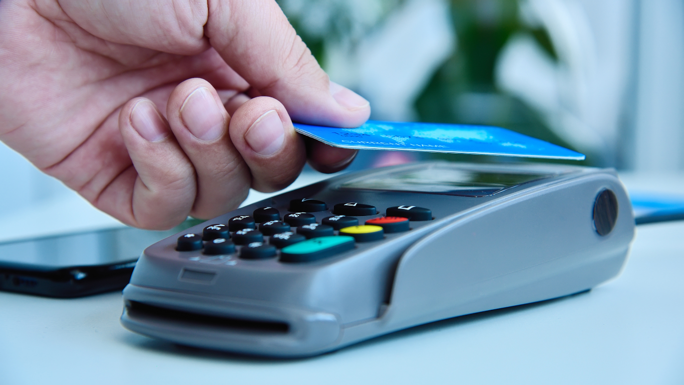 Close-up of a person's hand holding a blue credit card over a payment terminal, indicating a contactless payment in progress.